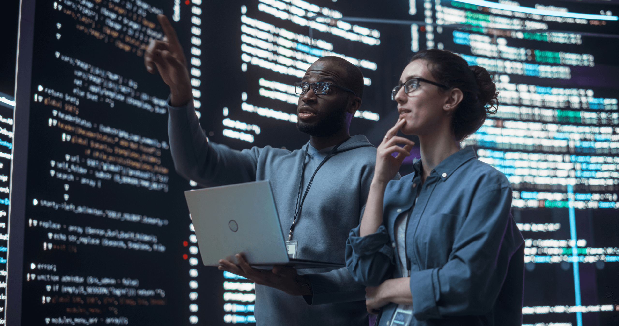 Two developers discussing lines of code appearing on big screens surrounding them